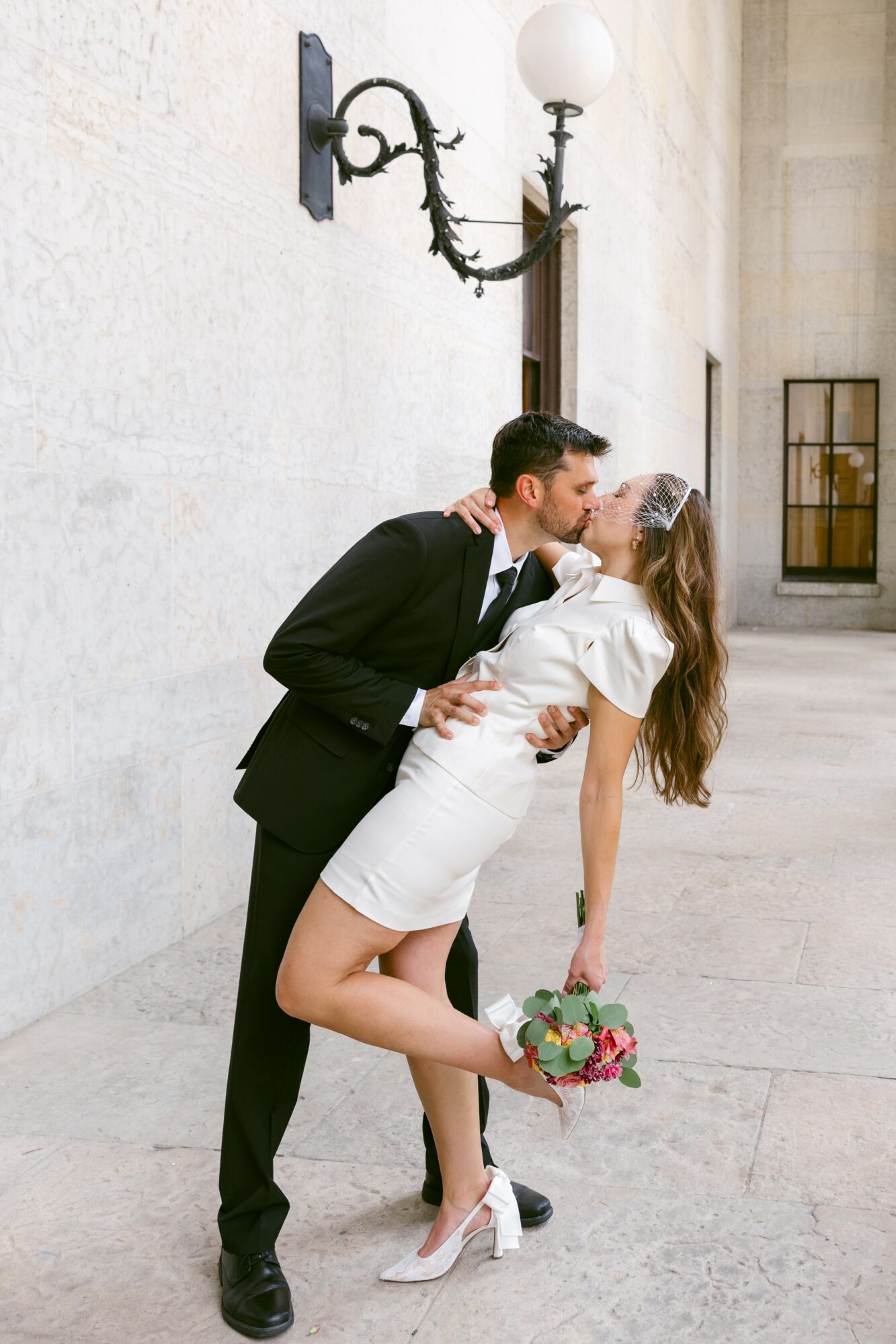 A couple in formal attire sharing a kiss outdoors, the woman holding a bouquet, against a plain wall with a lamp above.