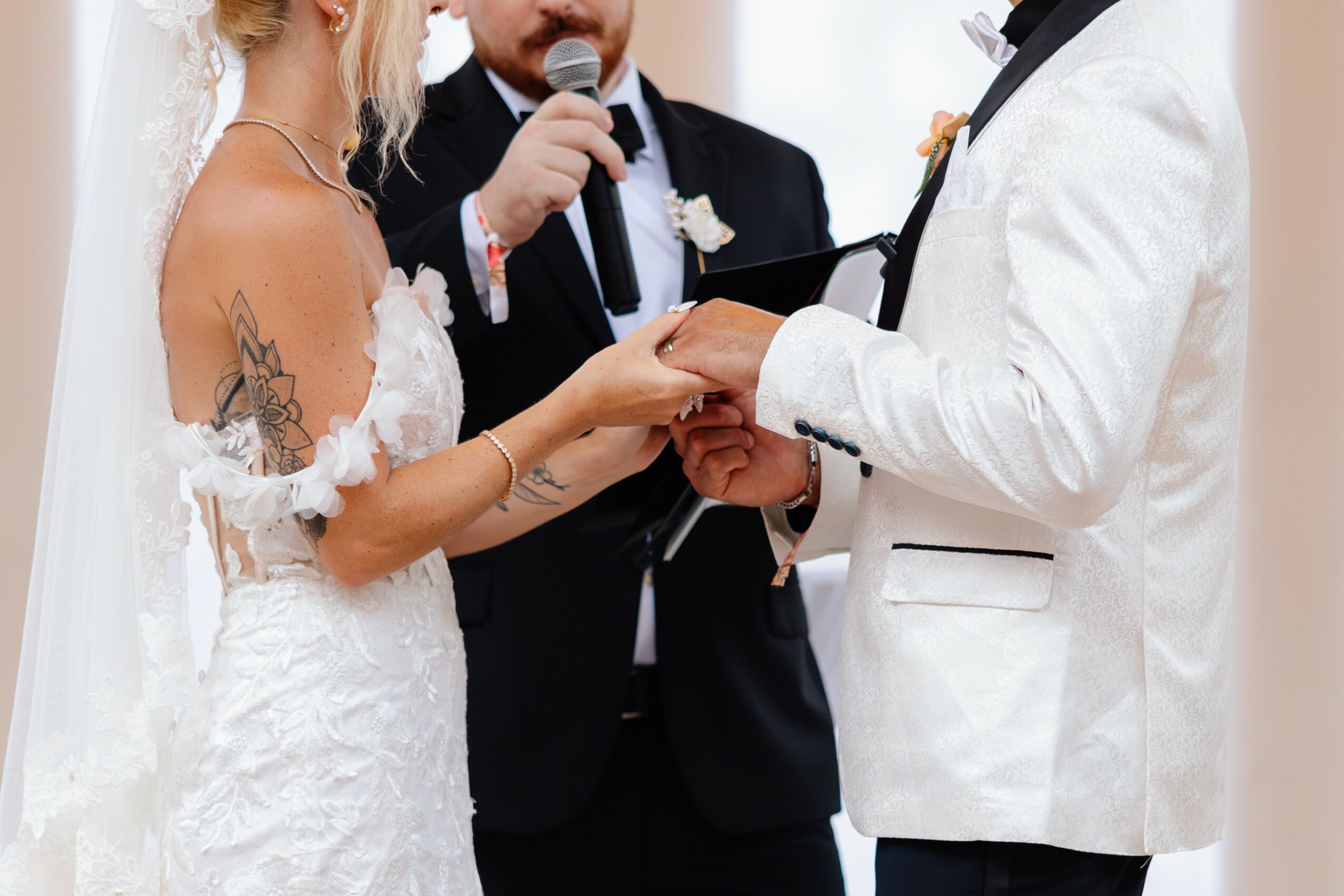 Bride and groom holding hands during wedding ceremony, officiant speaking in background, bride wearing white dress with tattoos, groom in white tuxedo