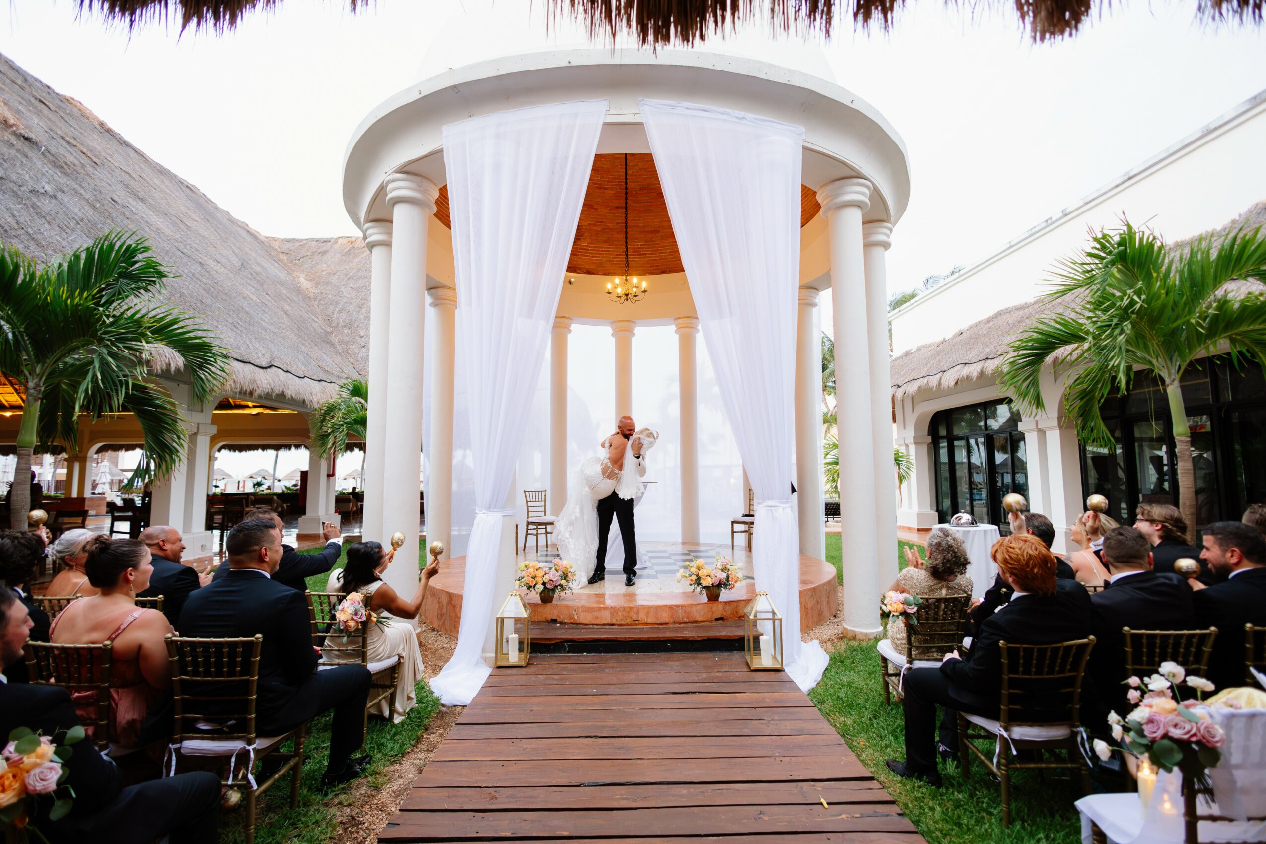 A wedding ceremony with a bride and groom on a wooden platform under a white circular pavilion, guests seated on both sides, tropical plants, and thatched roofs in the background.