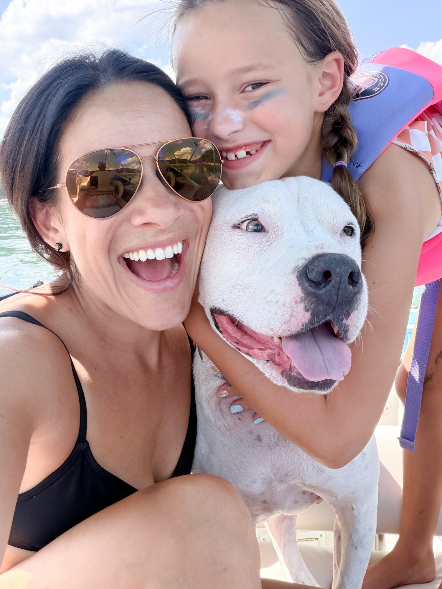 Two smiling females and a dog outdoors, with a blue sky and water in the background.