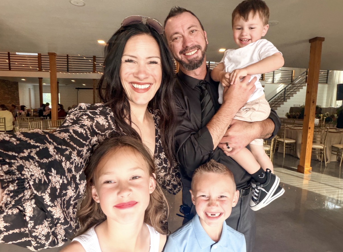 Family of four smiling indoors, woman taking selfie with man and two children, one girl and one boy, in background.