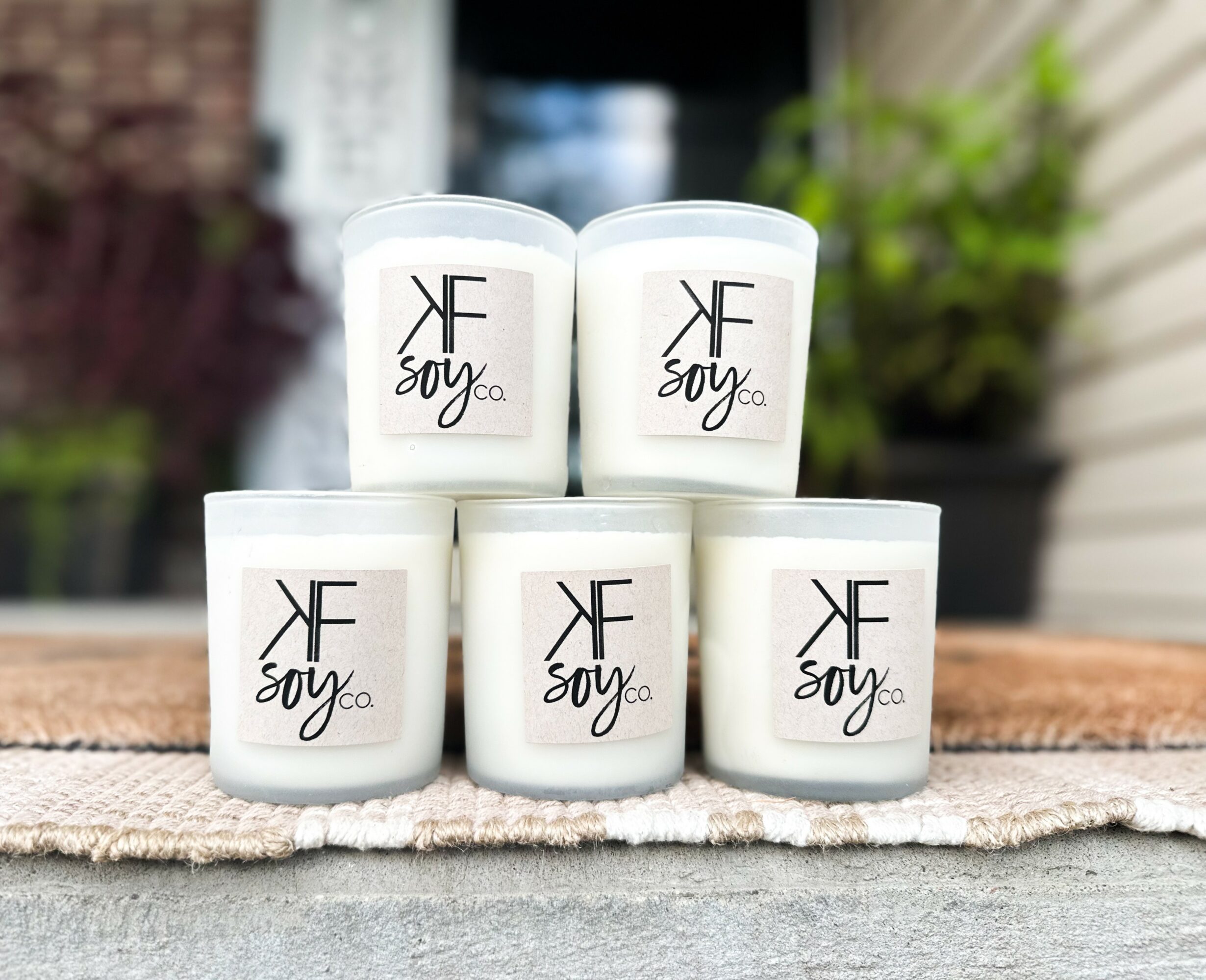 Five white containers with black text and logo, arranged outdoors on a wooden surface, with a blurred background of plants and building.