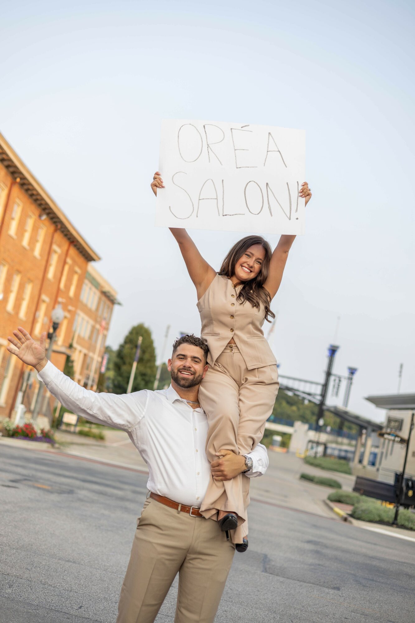 A man carries a woman on his shoulders holding a sign that reads 'Orea Salon' outdoors, with buildings and a street in the background.