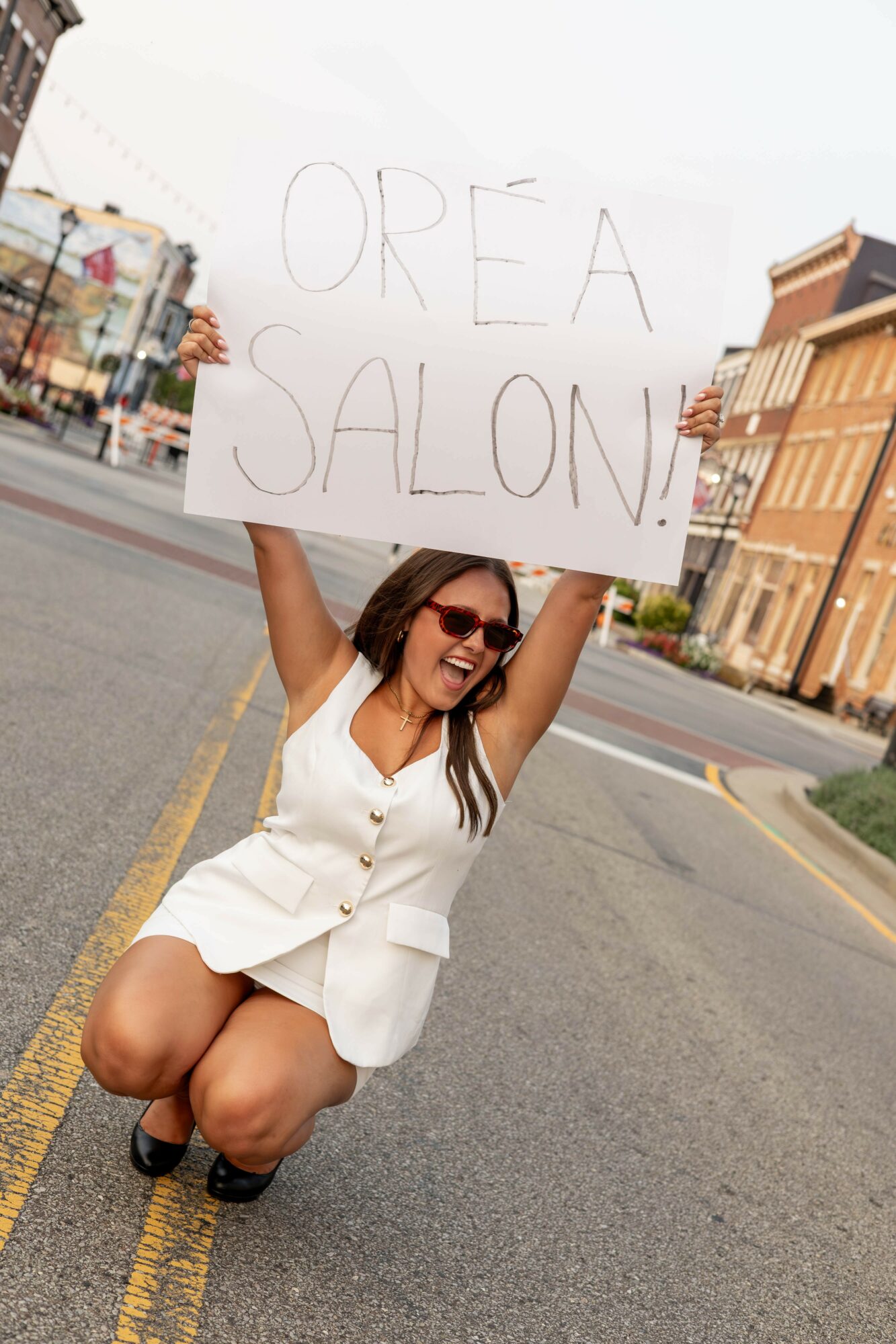 Woman kneeling on street holding a sign that says 'OREA SALON!', smiling, wearing sunglasses and a white outfit.