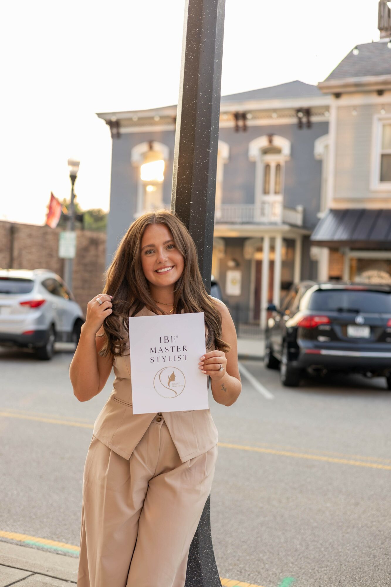 Young woman smiling holding a sign outdoors in front of houses and parked cars.