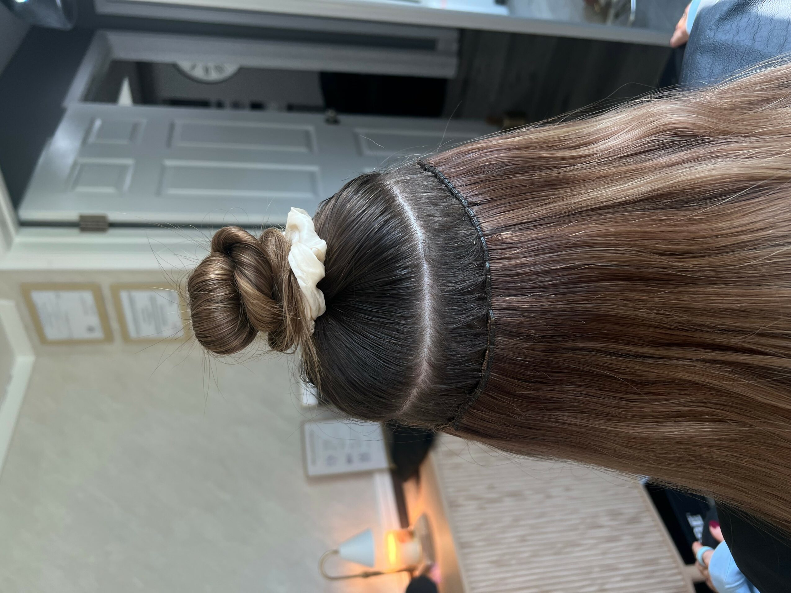 Back view of a person with long, straight brown hair tied in a bun with a white scrunchie, indoors.