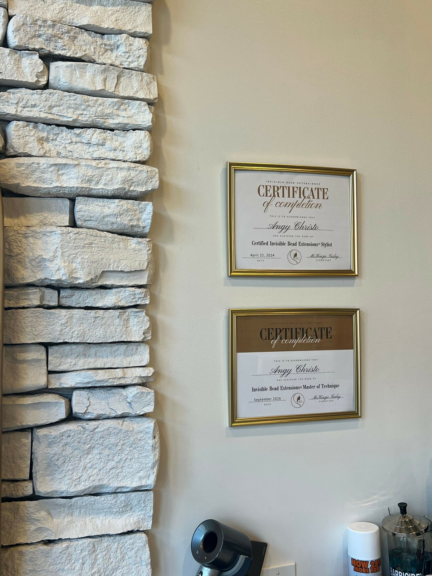 Two framed certificates hanging on a wall above a stone wall, with some bottles and objects on the right side.