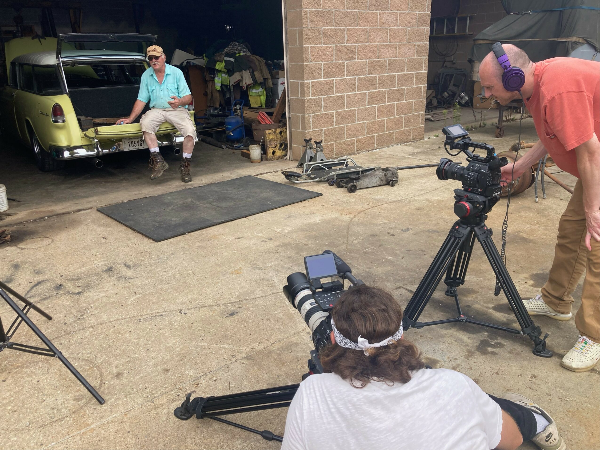 Person with headphones operating a camera on a tripod, filming a man sitting on a bench near a vintage vehicle, outdoors.