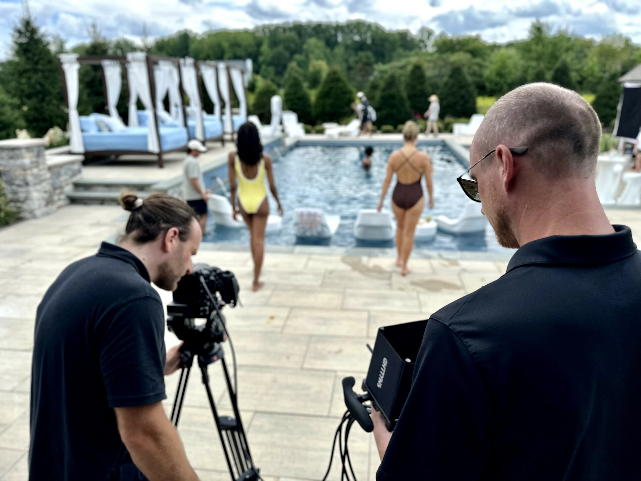Two men filming by a pool with people swimming and lounging, trees and umbrellas in the background.