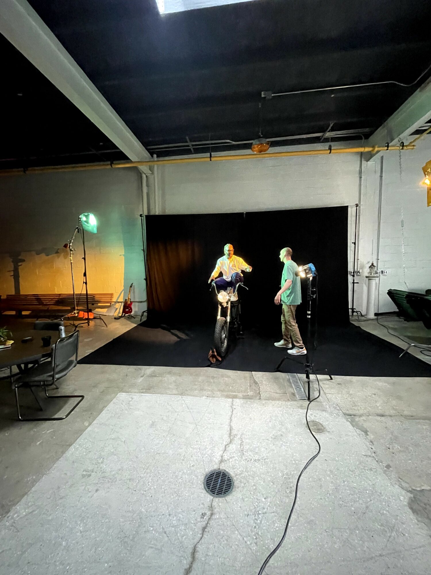 Two people on a stage with a black curtain, one playing guitar and the other standing, in an indoor venue.
