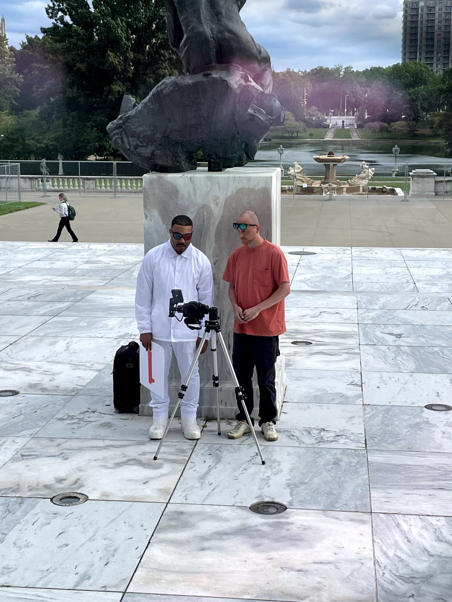 Two men stand in front of a large sculpture in a park, one with a camera on a tripod, the other with a backpack.