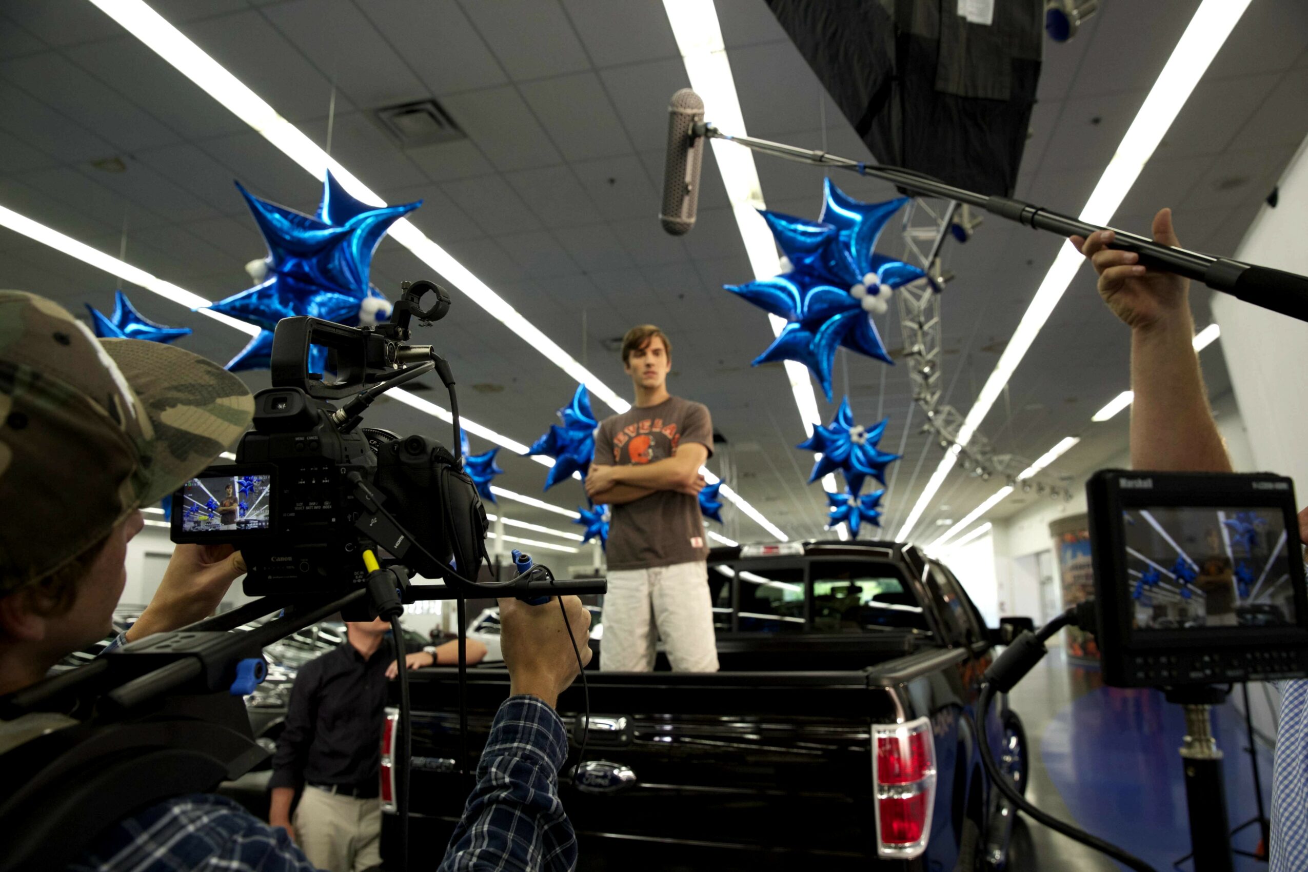 Person standing on a truck bed surrounded by blue star-shaped balloons, with cameras and crew filming in an indoor setting.