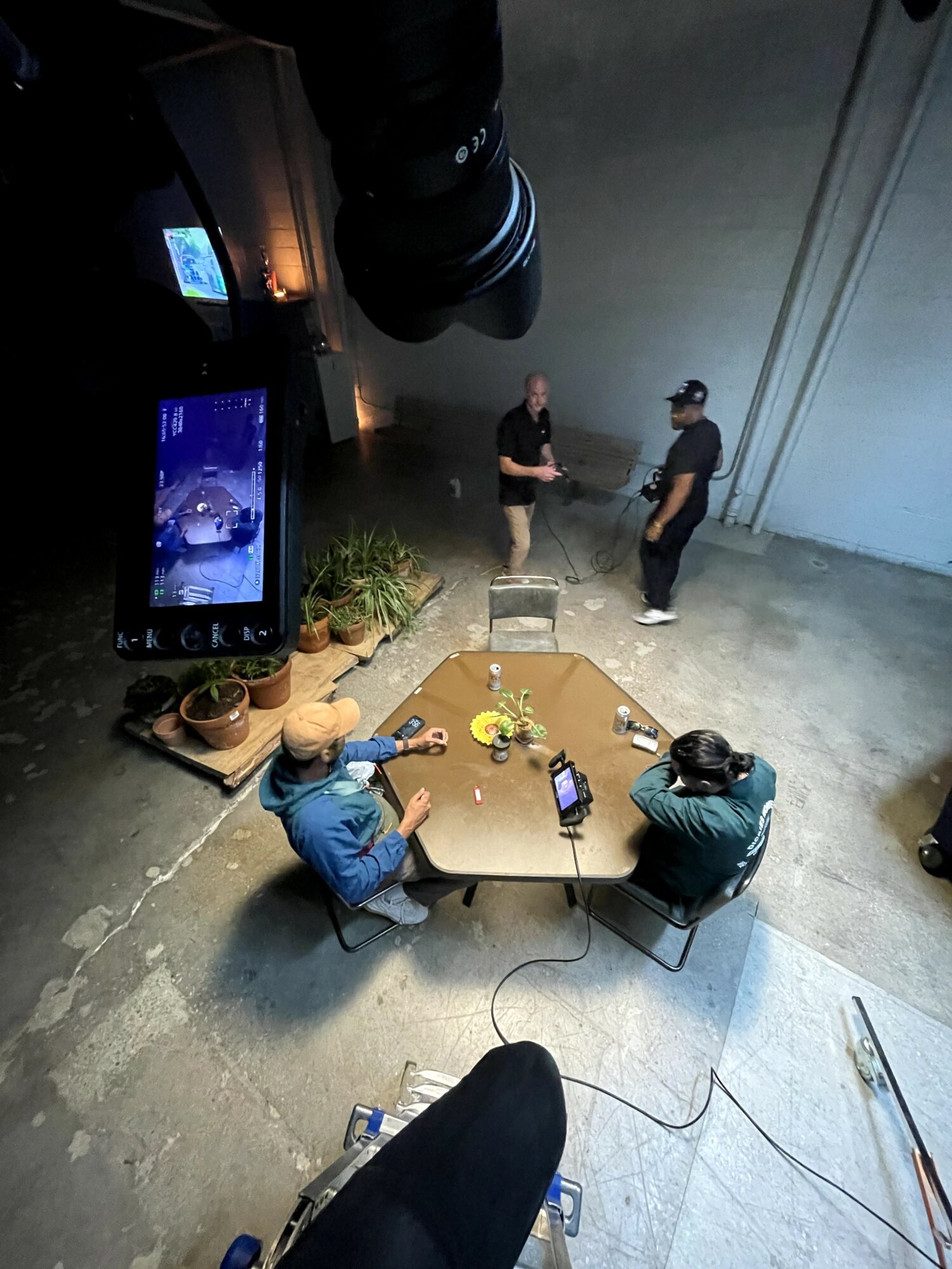 Two people sit at a table with cameras and equipment, two others stand and talk in a room with a large wall.