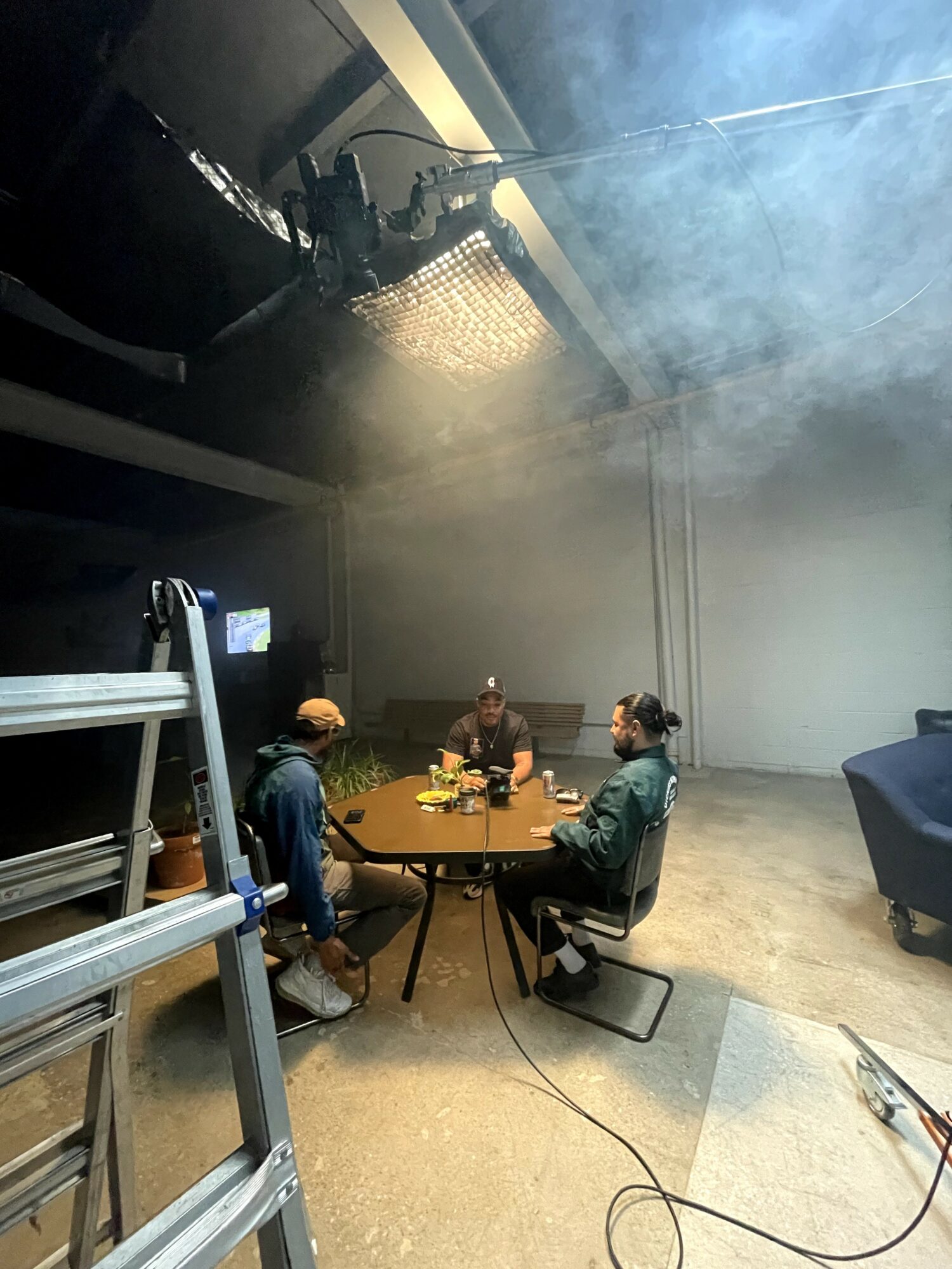 Four people sit around a table in a studio with smoke or fog, with equipment and a ladder nearby.