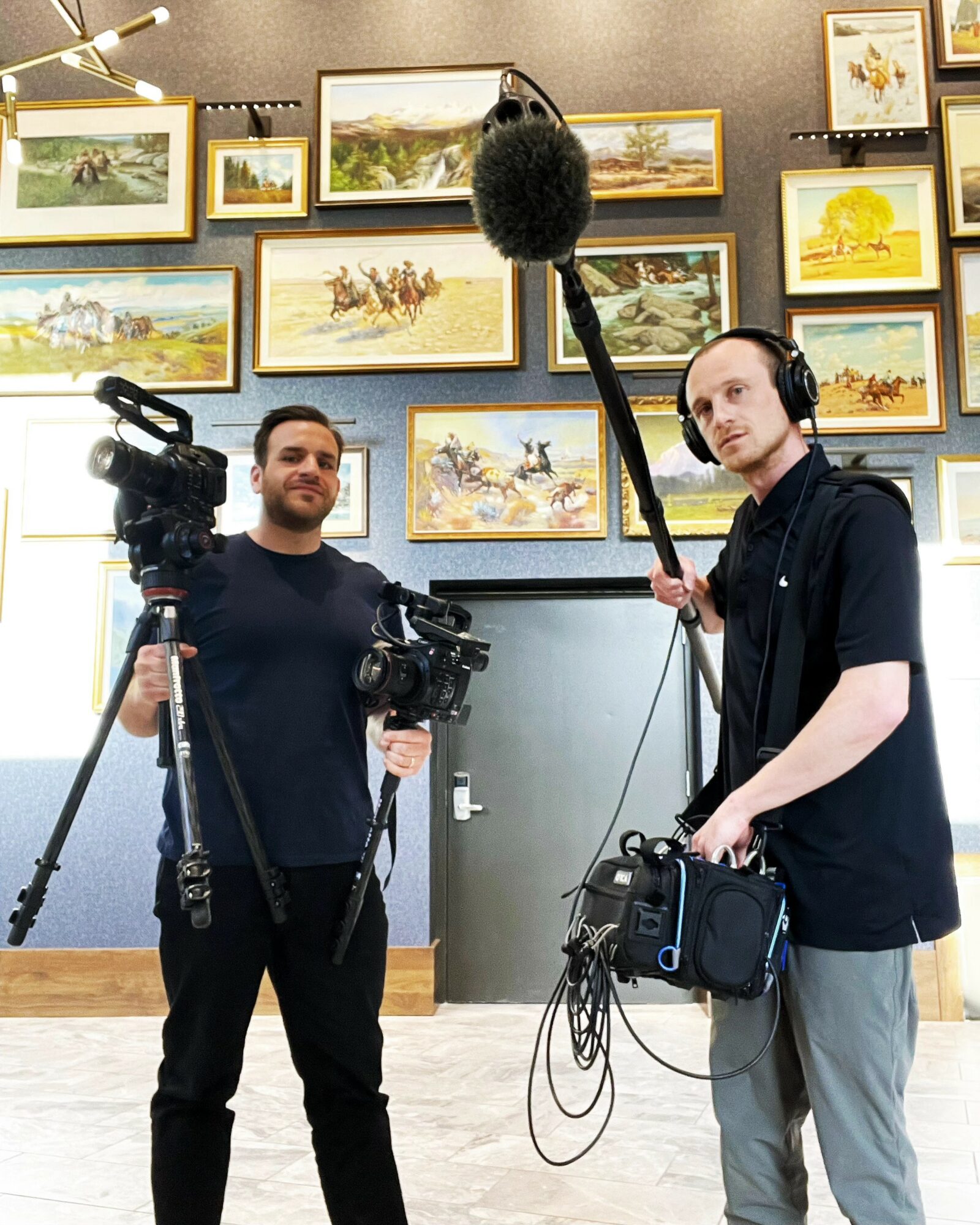 Two men with video equipment in an art gallery, one holding a camera and the other holding a boom microphone, in front of framed paintings.