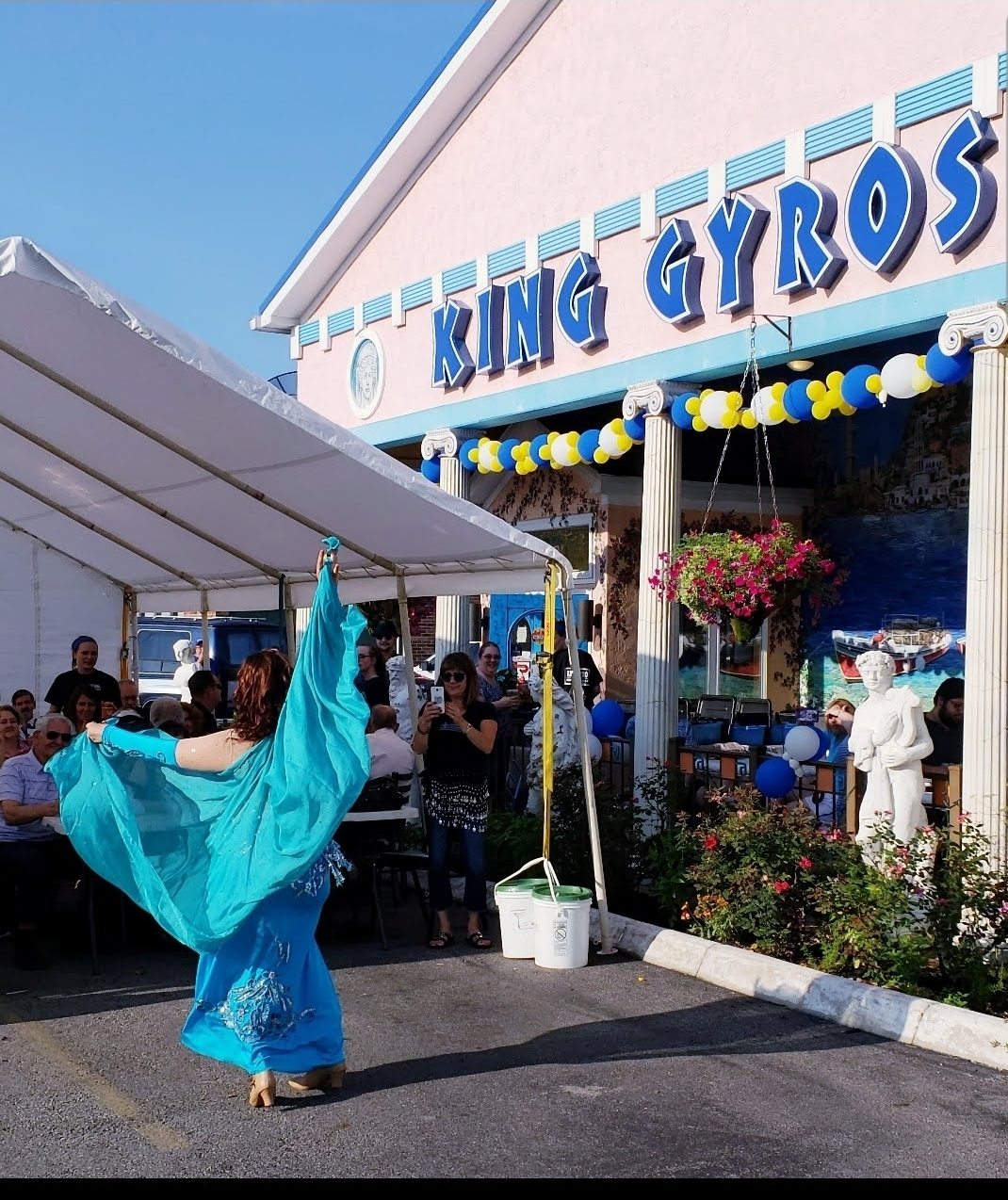 Person in blue costume performing a dance outside King Gyros restaurant, with people and decorations nearby.