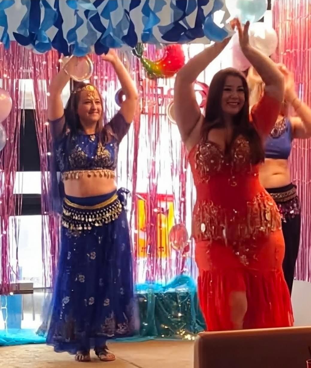 Two women in colorful dresses dance with arms raised, decorated with hanging streamers and balloons in the background.