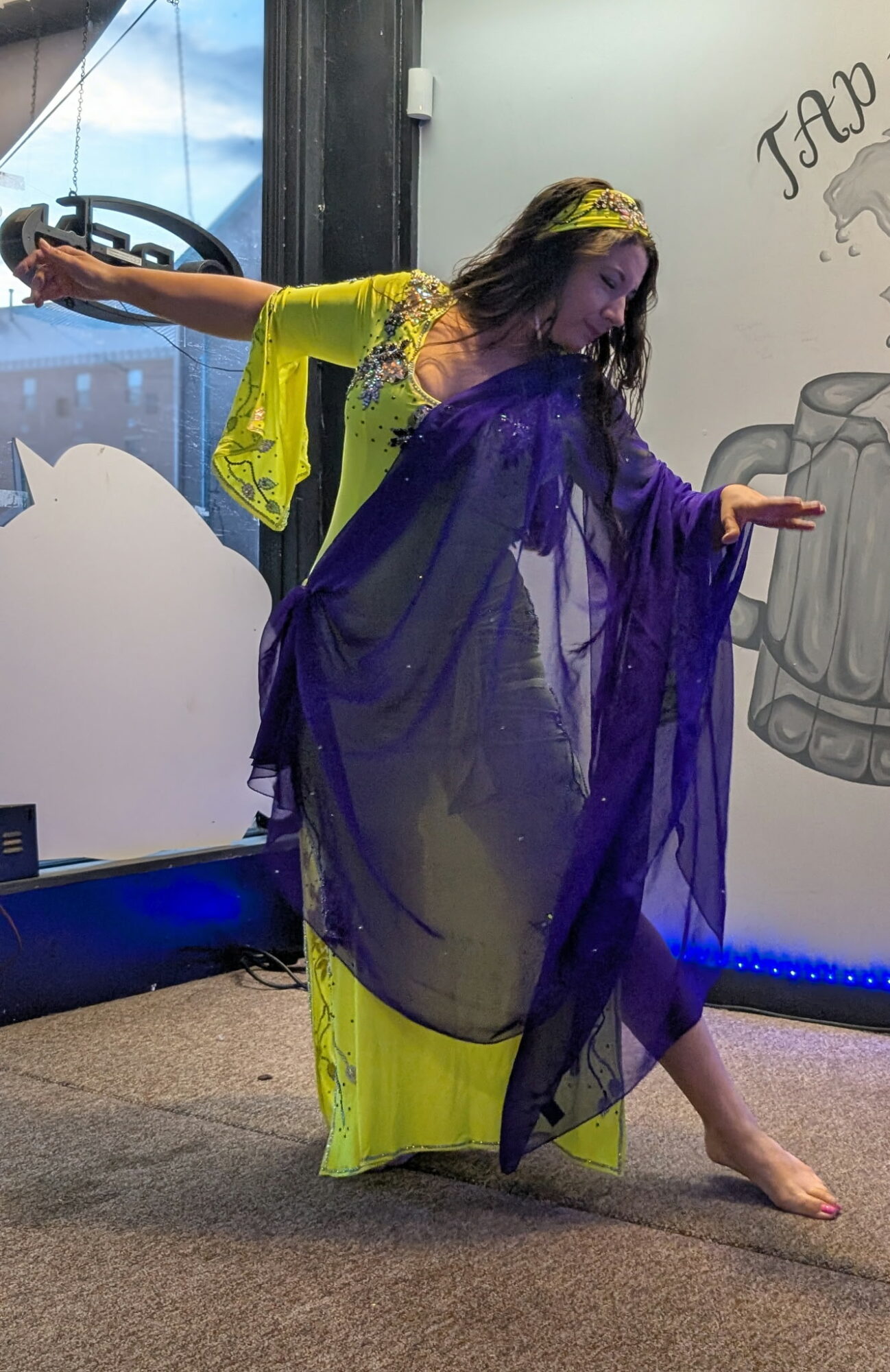 Person in colorful costume poses with one arm extended and one leg bent, standing on a stage with a painted backdrop.