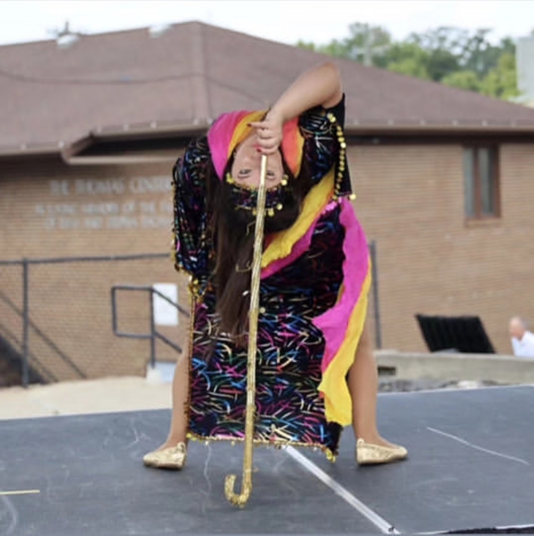 Child in colorful costume bending forward holding a stick, on a stage outdoors with a building in background.