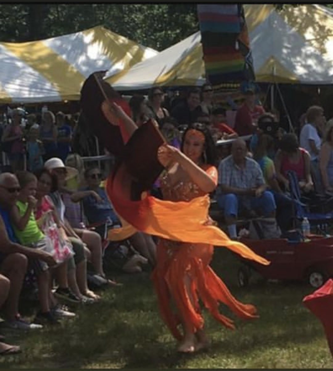 Person performing a dance in an orange costume outdoors with an audience and tents in background.