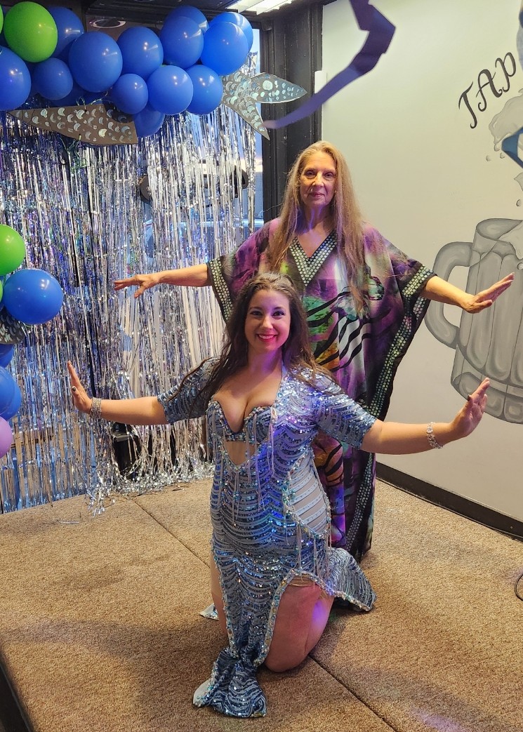 Two women in colorful costumes pose with arms outstretched in front of a decorated backdrop with balloons and streamers.