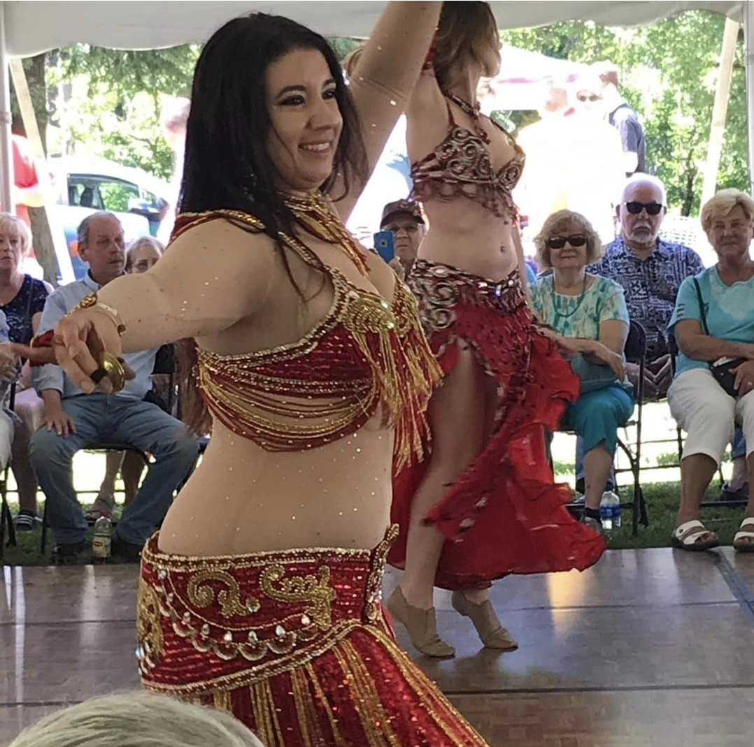 Two belly dancers perform outdoors with an audience watching, one smiling and wearing a red and gold costume, the other in a patterned outfit.