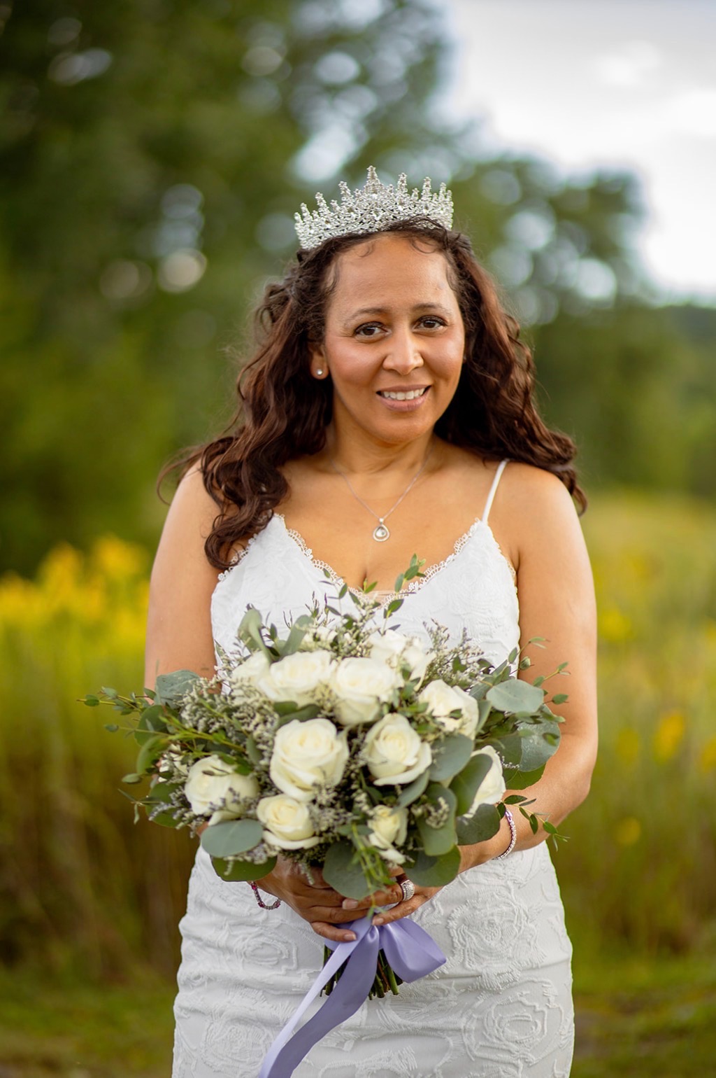 Woman in wedding dress holding bouquet outdoors, wearing a tiara, smiling, with trees and sky in background.