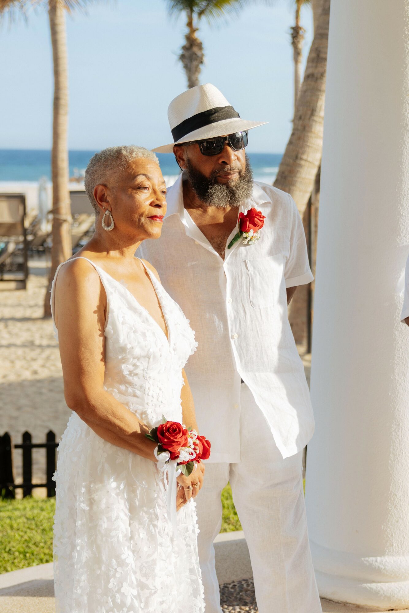 An elderly woman and a man stand outdoors near white columns, with palm trees and the ocean in the background.