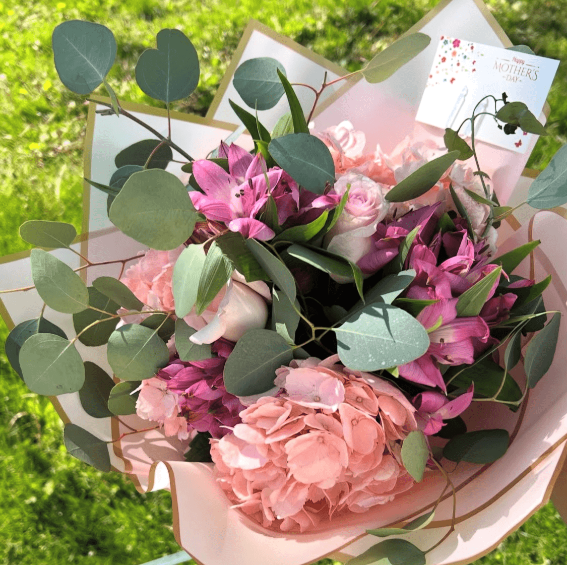 Bouquet of pink and purple flowers with green leaves, wrapped in light pink paper, outdoors with green foliage background.