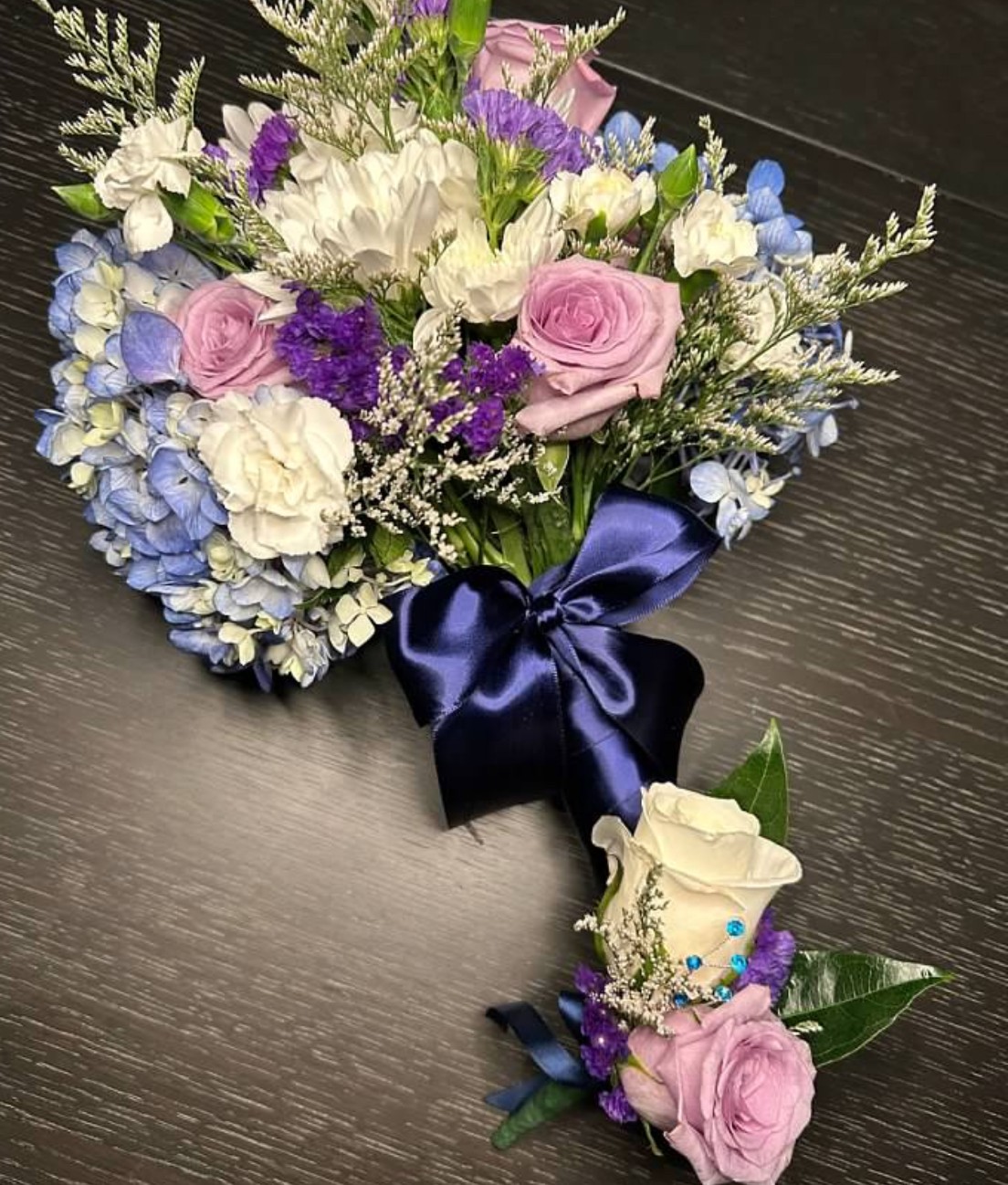 Flower arrangement with pink, white, purple, and blue flowers, tied with a dark blue ribbon, on a dark surface.