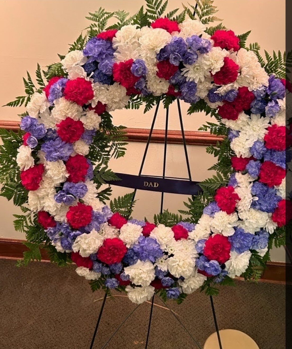 A floral wreath with red, white, and purple flowers on a stand, with a black ribbon reading 'DAD' in the center.