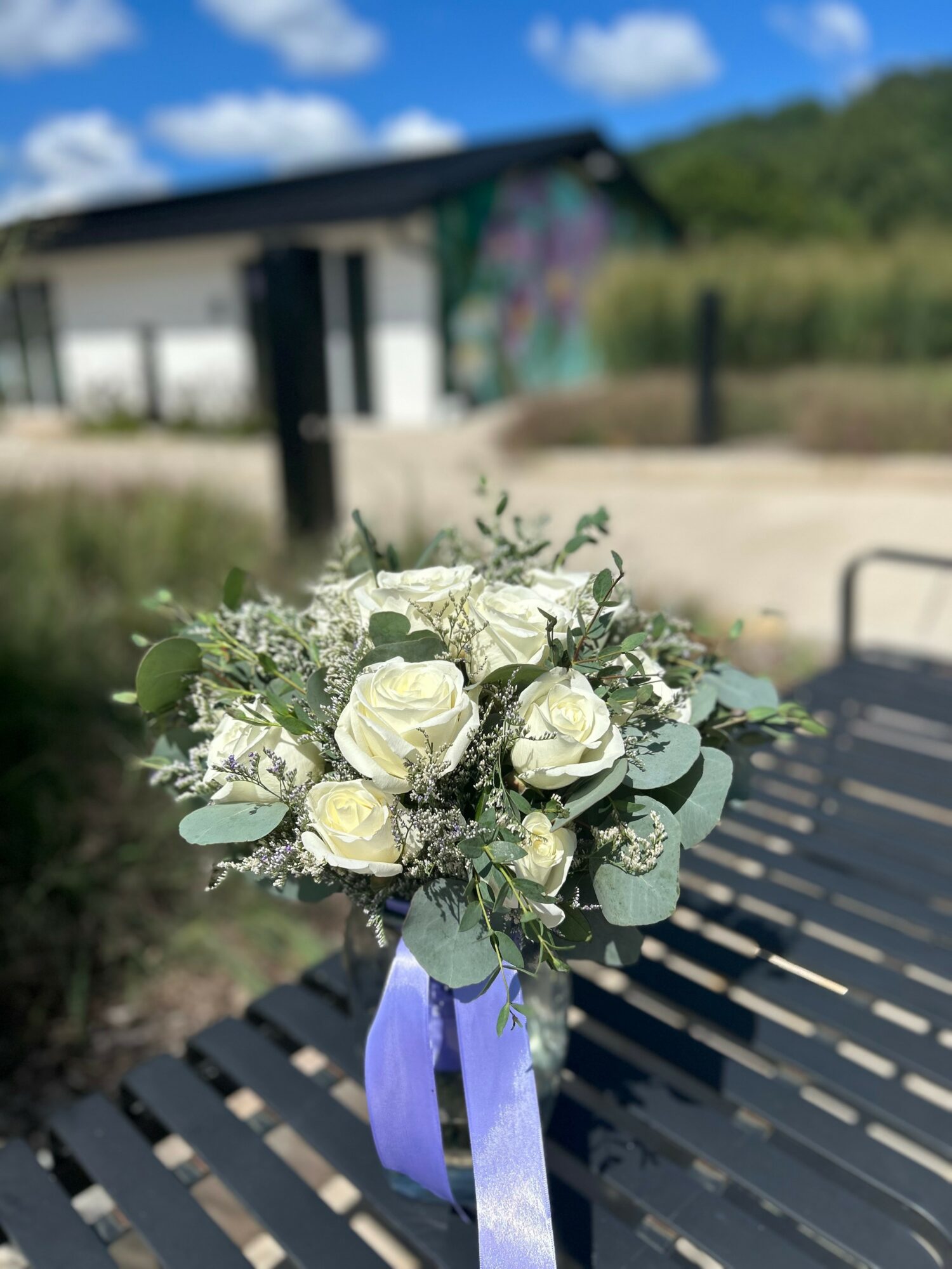 Bouquet of white roses and greenery with a purple ribbon, placed on a black metal bench outdoors.