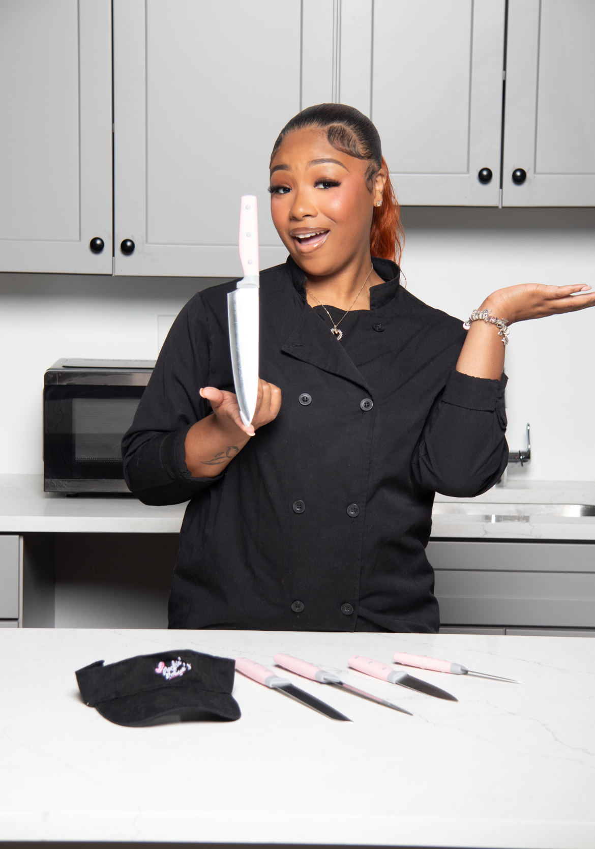 Woman in chef's coat holding a knife, smiling in a kitchen with gray cabinets, with knives and a cloth on the counter.