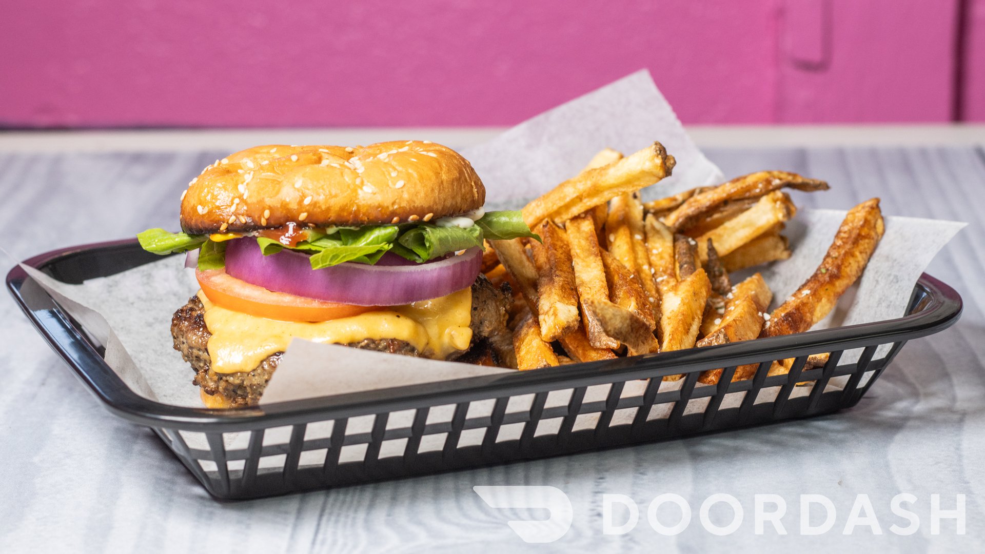 Burger with lettuce, tomato, onion, cheese, and a bun, served with French fries in a black basket on a gray surface.
