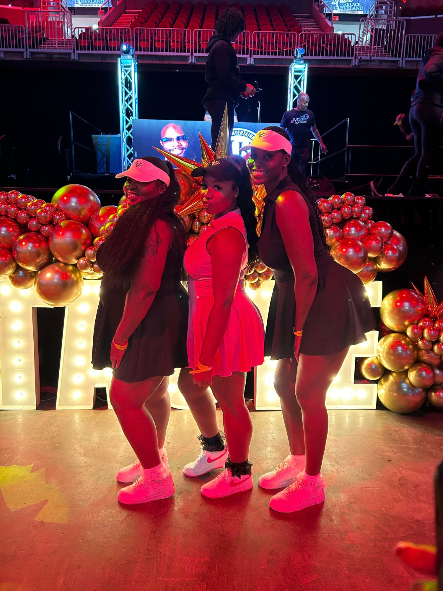 Three people standing together in a festive setting with balloons and bright lights, wearing casual clothing and pink shoes.