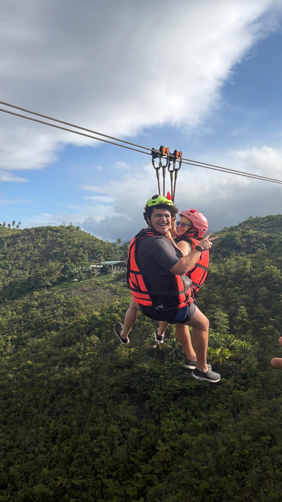 Person and child riding a zip line over a green landscape with hills and a partly cloudy sky.