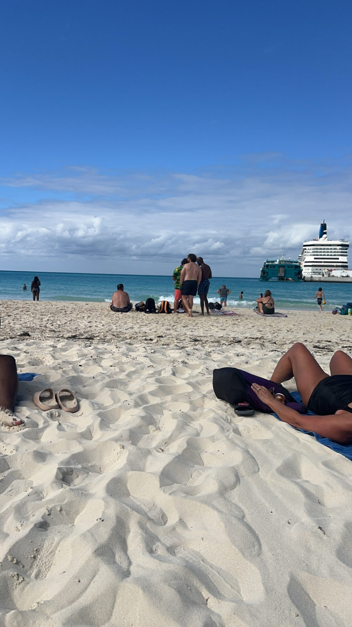 People on a sandy beach with a large cruise ship in the ocean, blue sky, and some clouds.