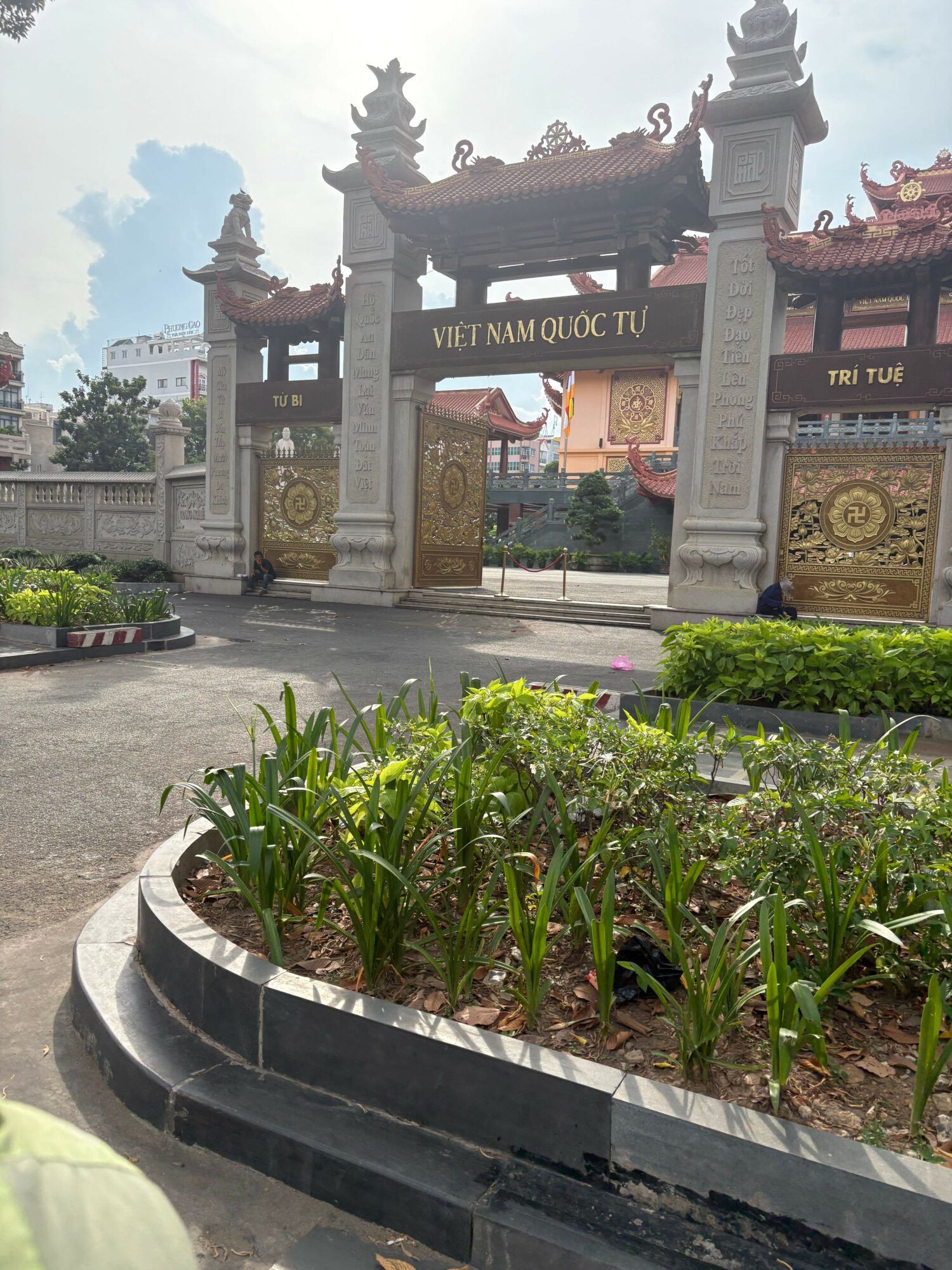 Street scene with decorative archway, plants, and buildings in the background, viewed from the side.