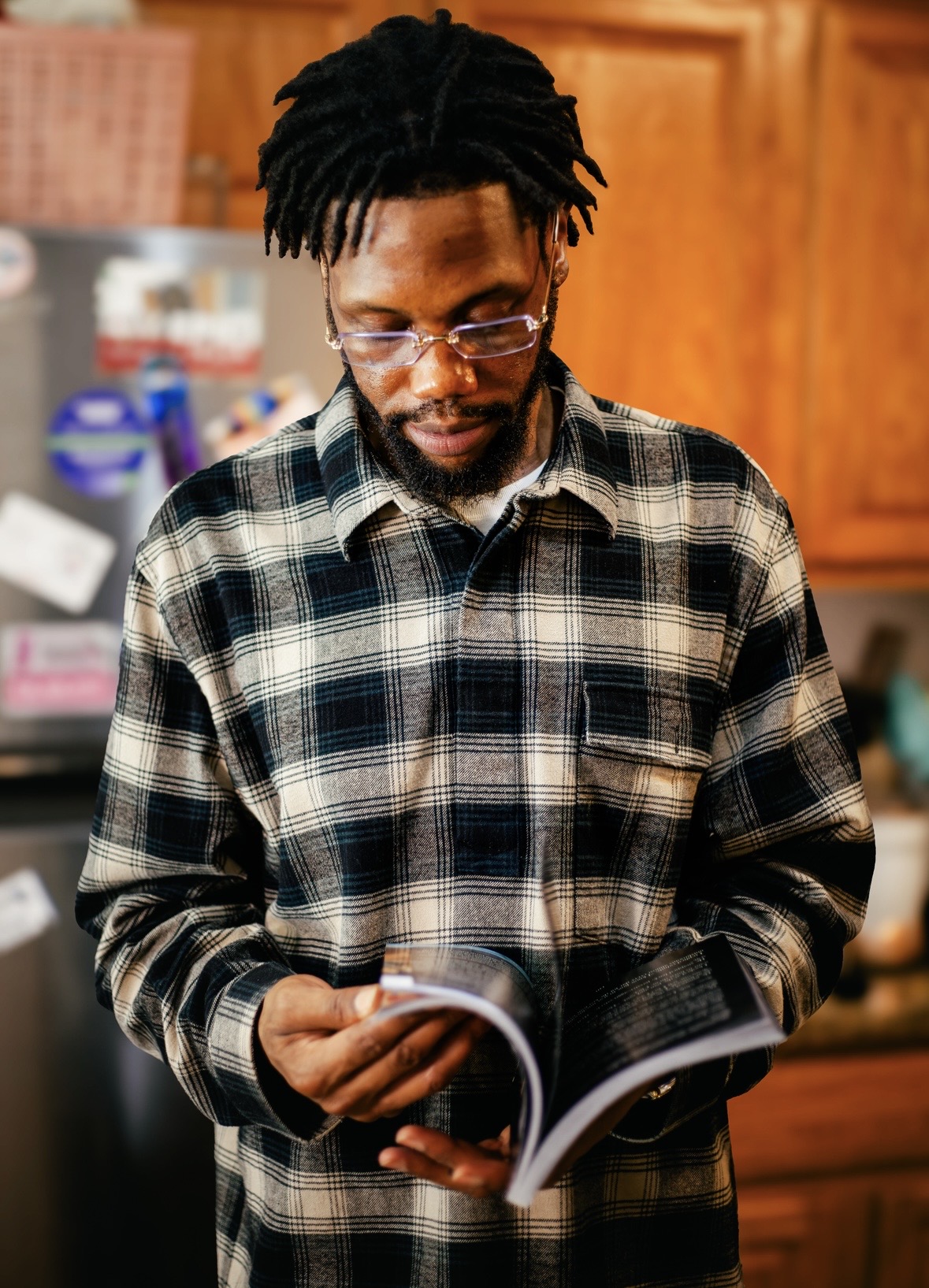 Man with glasses and dreadlocks reading a book in a kitchen with wooden cabinets.