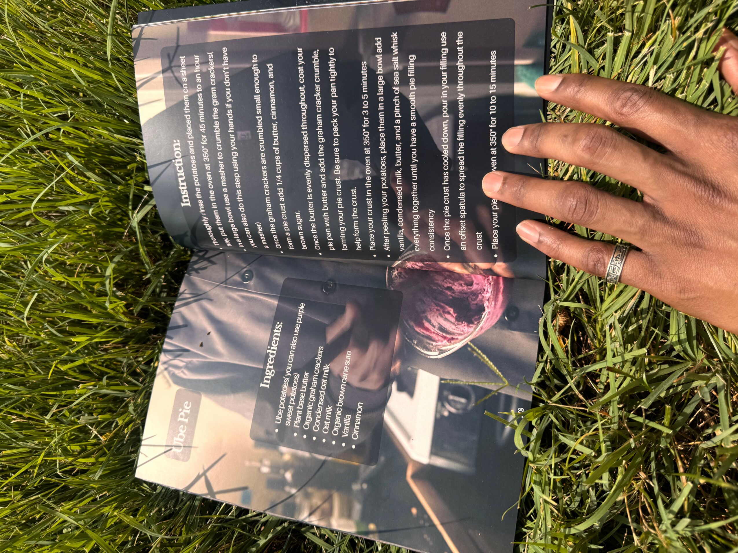 Person's hand holding an open book with text and an image, on grass outdoors.