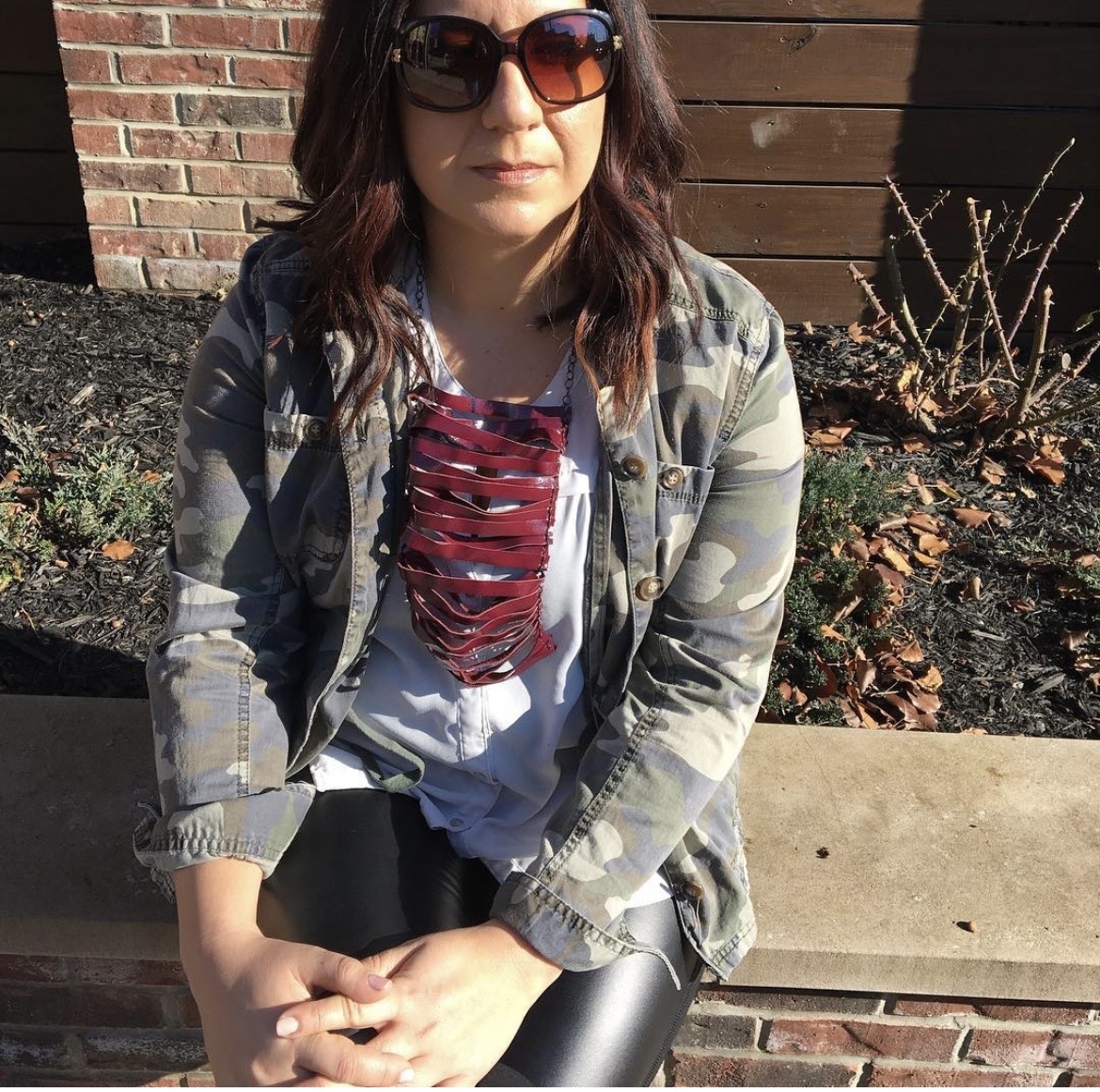 Woman with sunglasses sitting outdoors on a bench, wearing a camouflage jacket and a distressed red and white shirt.