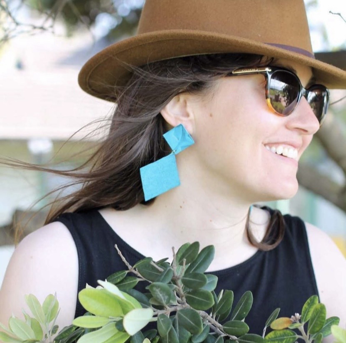 Woman wearing sunglasses, a hat, and blue earrings, holding a bouquet of green plants, smiling outdoors.
