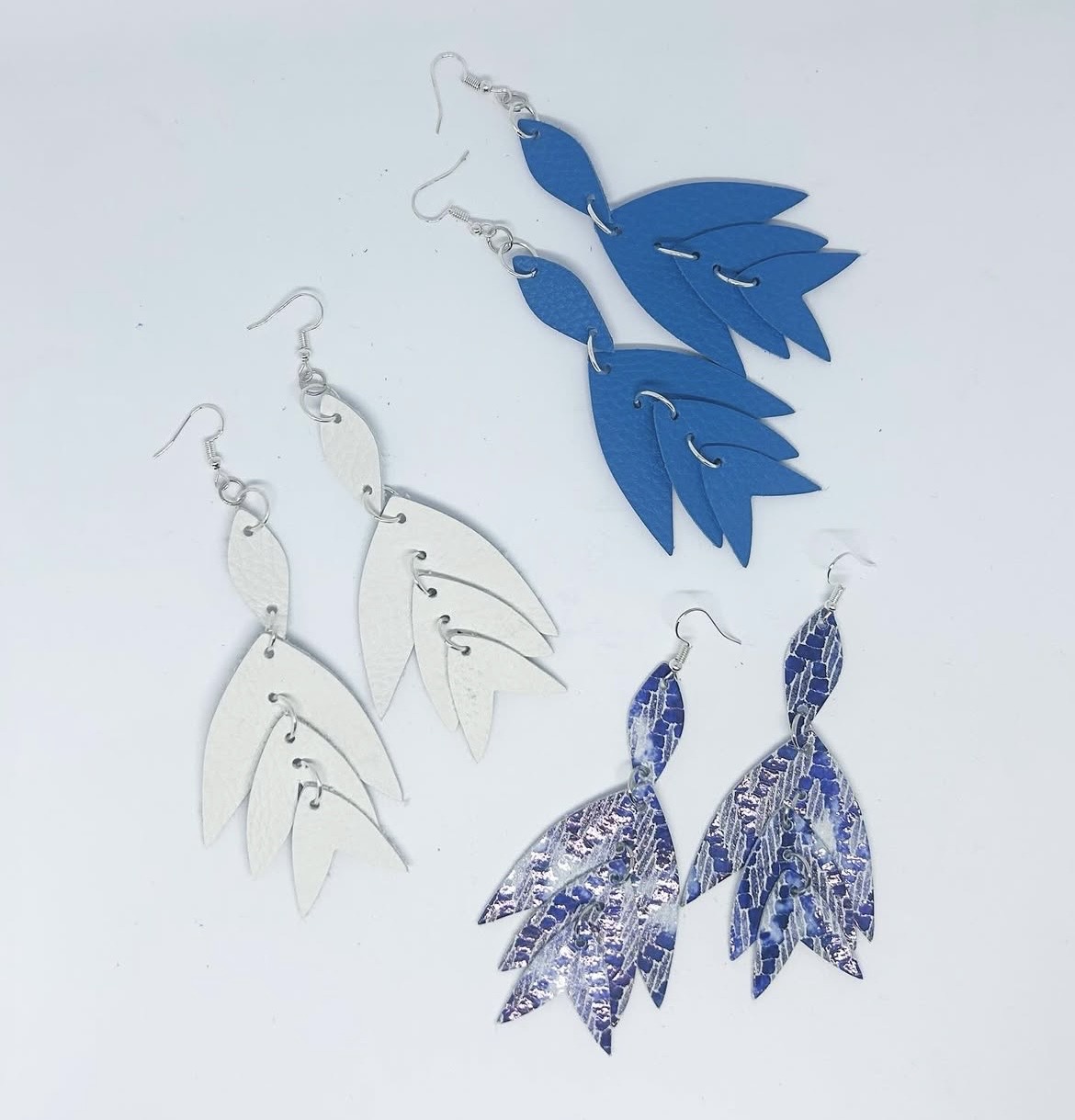 Three pairs of leaf-shaped earrings in blue, white, and patterned designs, arranged on a light background.