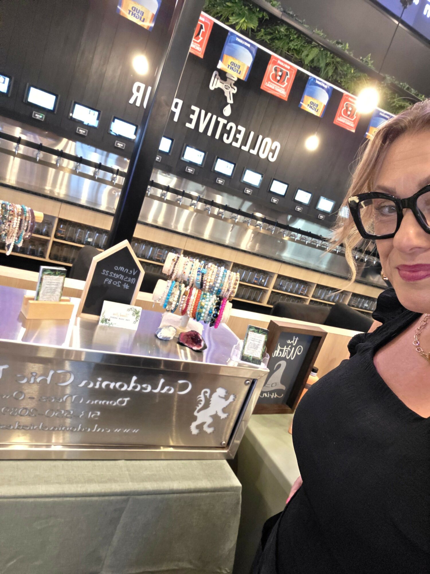 Woman with glasses at a counter with colorful items and a sign, in a store or cafe setting.