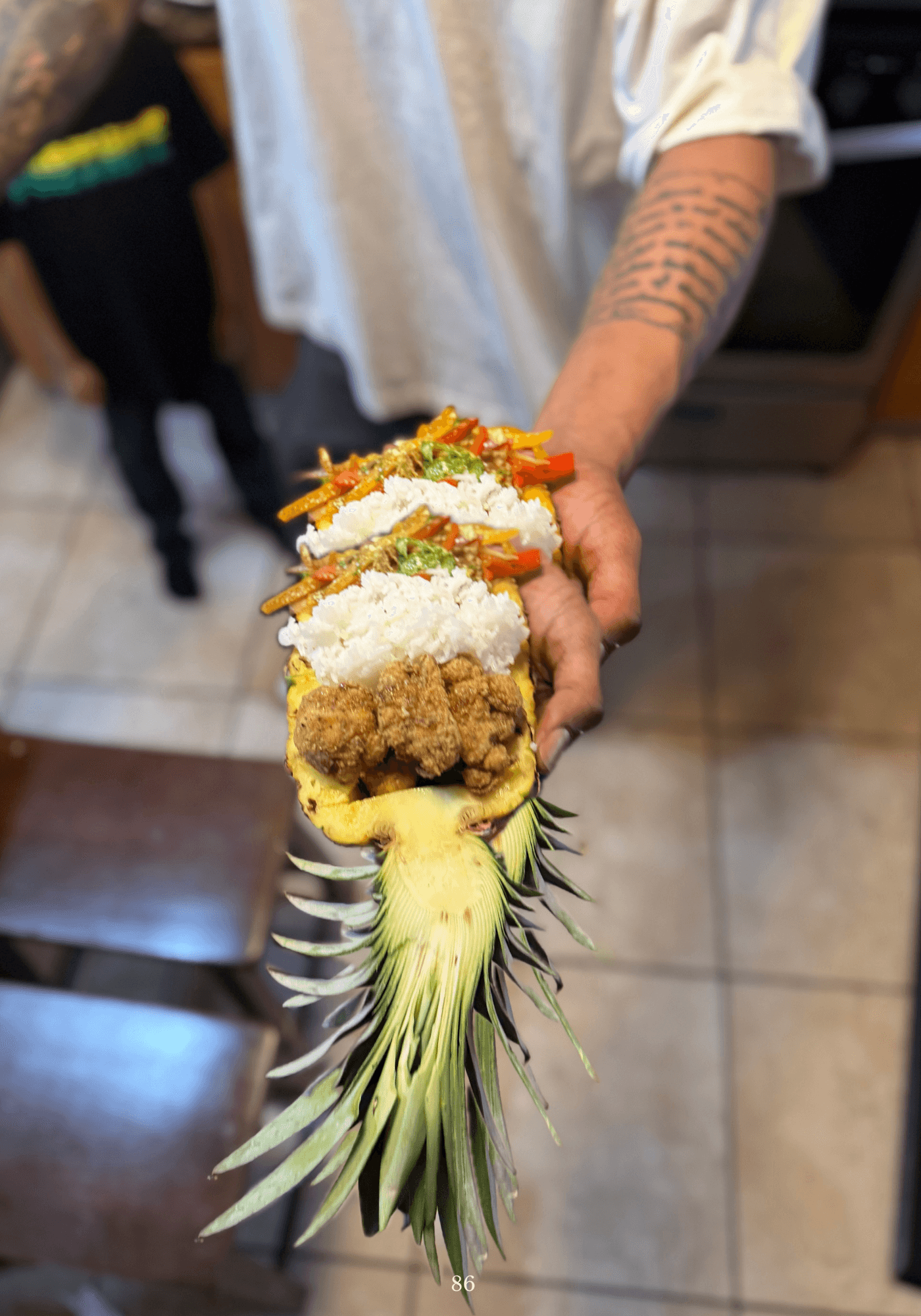 Person holding a pineapple boat filled with rice, fried chicken, vegetables, and toppings, in a kitchen setting.