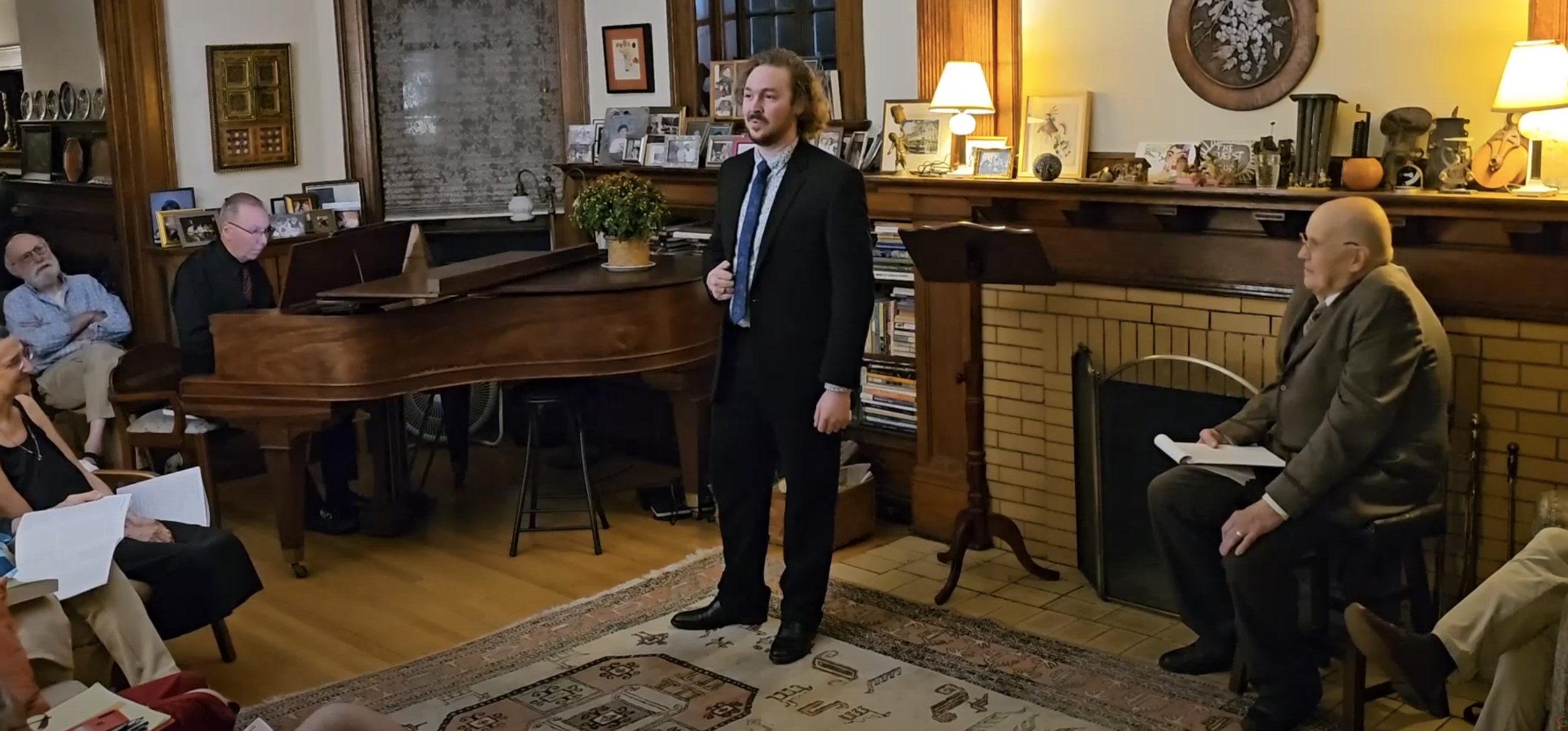 Man in suit standing in a room with people seated, piano, and wooden furniture, speaking to an audience.