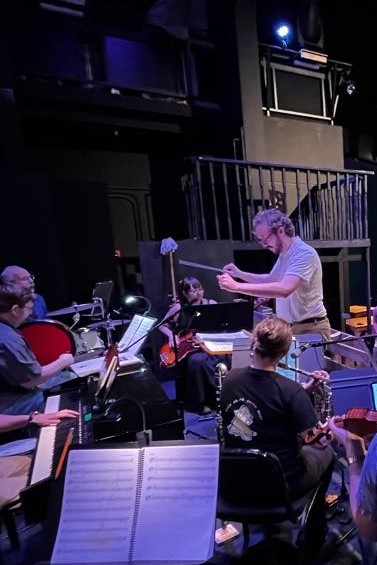 Orchestra musicians playing instruments on stage with sheet music, stage lighting, and a dark background.
