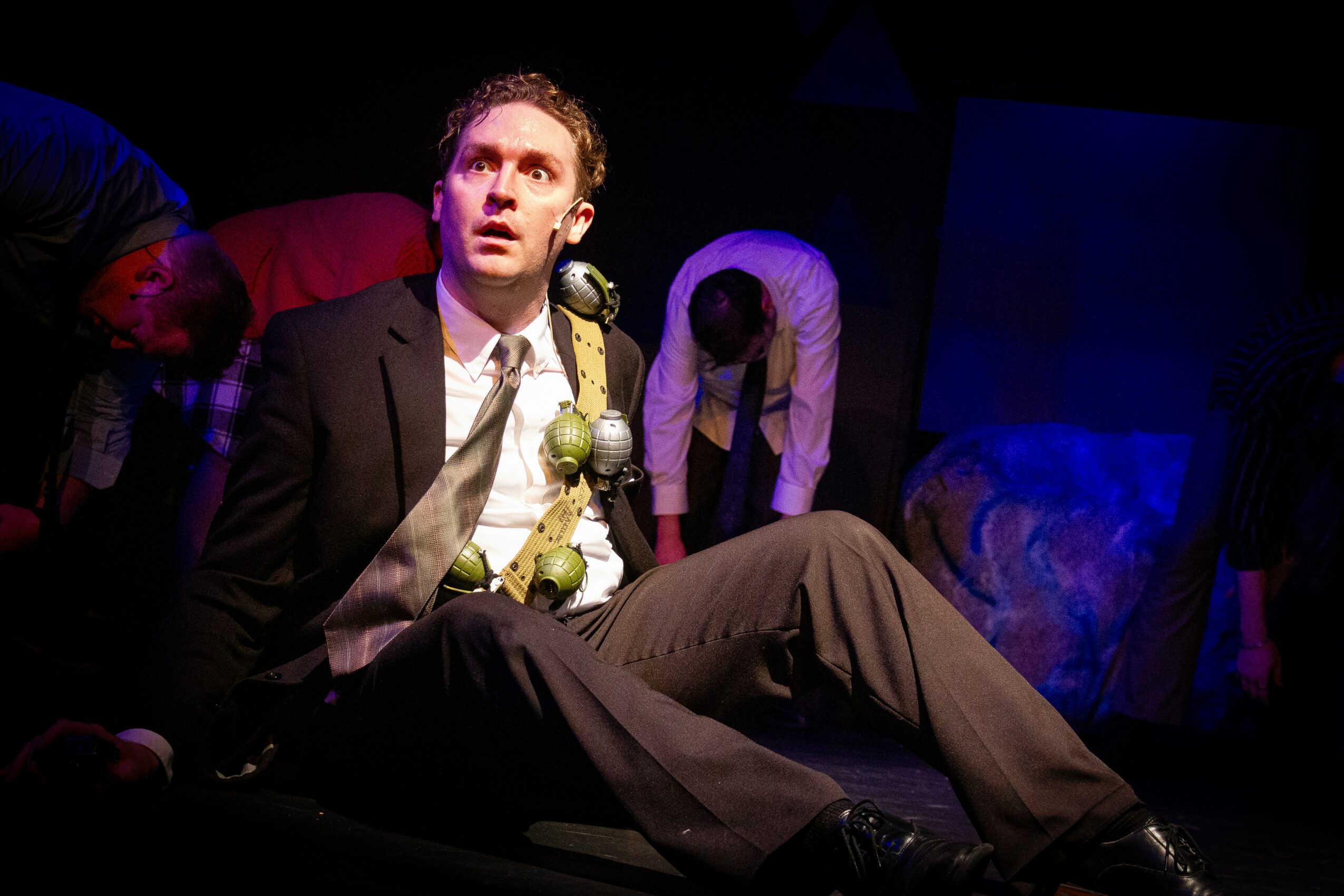 Man in a suit sitting on the floor with a surprised expression, surrounded by people in a dark setting.