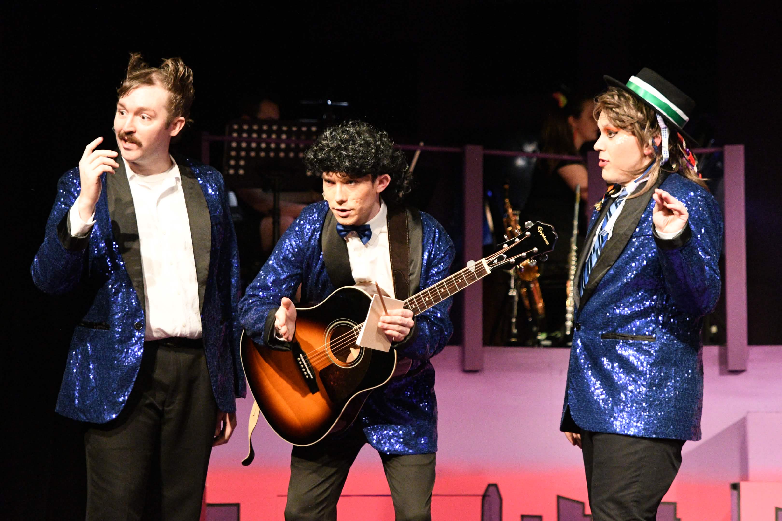 Three young men in blue sequined jackets perform on stage, one playing guitar, with a pink backdrop and city lights in background.