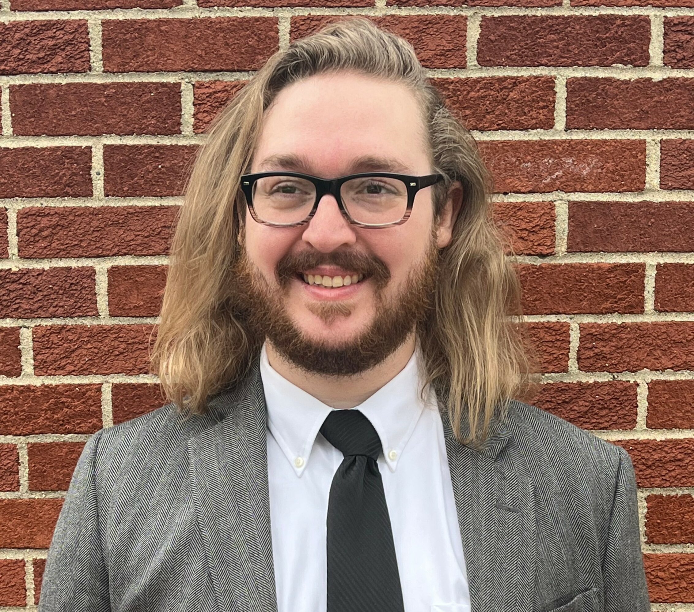 Man with long hair, beard, glasses, wearing a suit and tie, smiling in front of a brick wall.